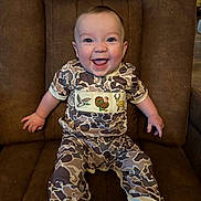 Walker joined the competition — help win amazing prizes! baby, child, smiling, camo_clothing, chair, brown_furniture, happy, sitting, cute, infant, portrait, indoors, person, young_child, embroidered_clothing, cozy, comfortable, bright_eyes, small_hands, soft_lighting