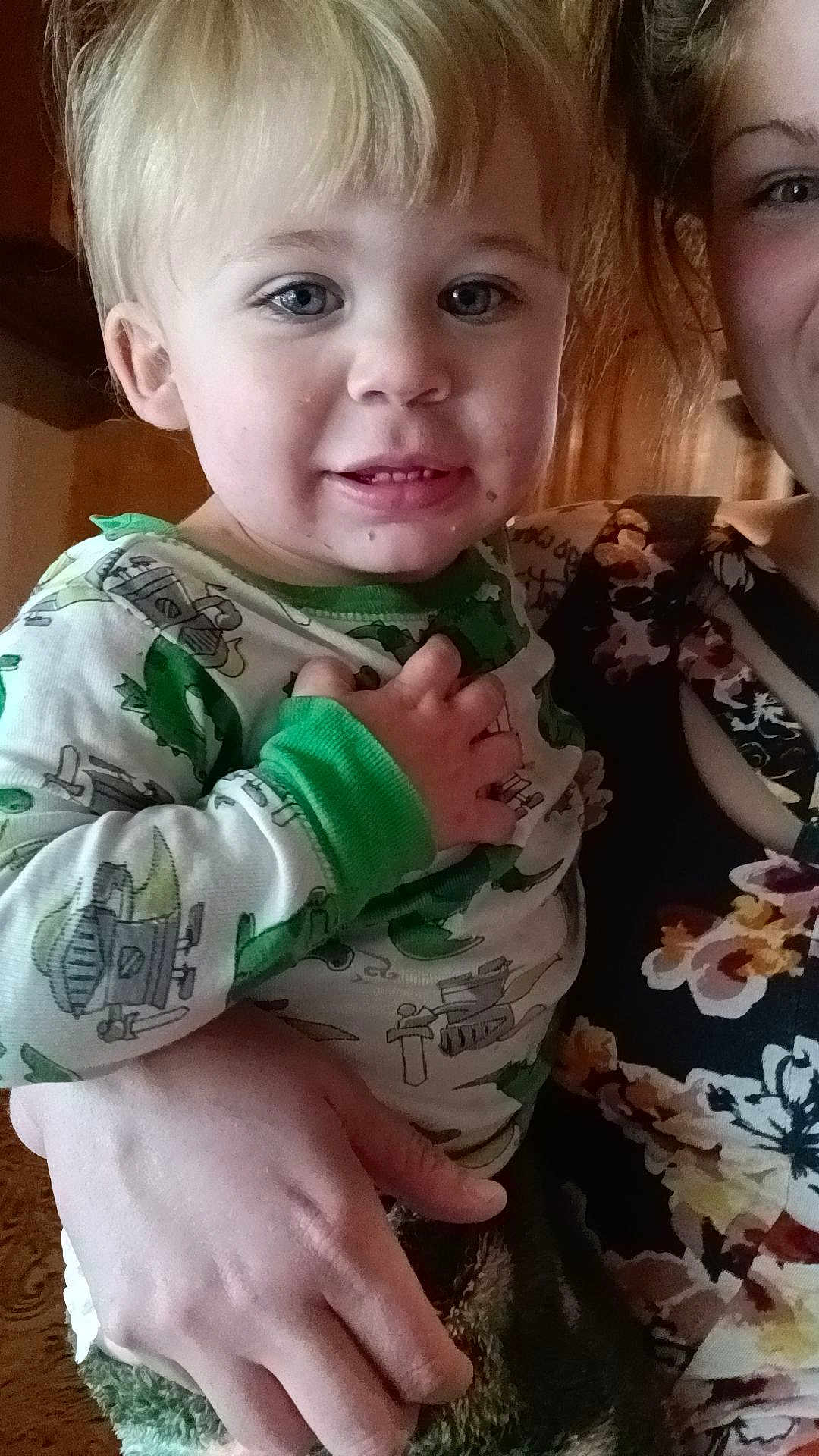 Asher is registered to the contest to win money with this photo: child, toddler, person, face, blonde_hair, blue_eyes, pajamas, green_clothing, dinosaur_pattern, hand, adult, floral_clothing, indoor, portrait, smile, curious, holding, closeup, skin, hair