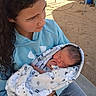 child, baby, newborn, blanket, outdoor, person, holding, curly_hair, hoodie, blue_clothing, sleeping, infant, young_girl, casual_clothing, vehicle, dirt_ground, tender_moment, wrapped, peaceful, family
