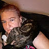 person, dog, brindle_dog, pet, cuddling, indoors, couch, portrait, selfie, closeup, face, eyes, blonde_hair, black_clothing, patterned_shirt, pink, relaxed, affection, companion, home