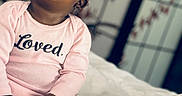 Kailani is registered to the contest to win money with this photo: baby, child, infant, pink_onesie, clothing, face, hands, bed, white_bedspread, indoor, portrait, sitting, looking_up, curious, expression, skin, person, young_child, cute, soft_light