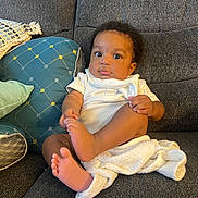 Jaxson is registered to the contest to win money with this photo: baby, child, infant, couch, pillow, blanket, white_clothing, curly_hair, indoor, cute, sitting, hand, foot, face, expression, comfort, home, soft_texture, relaxed, portrait