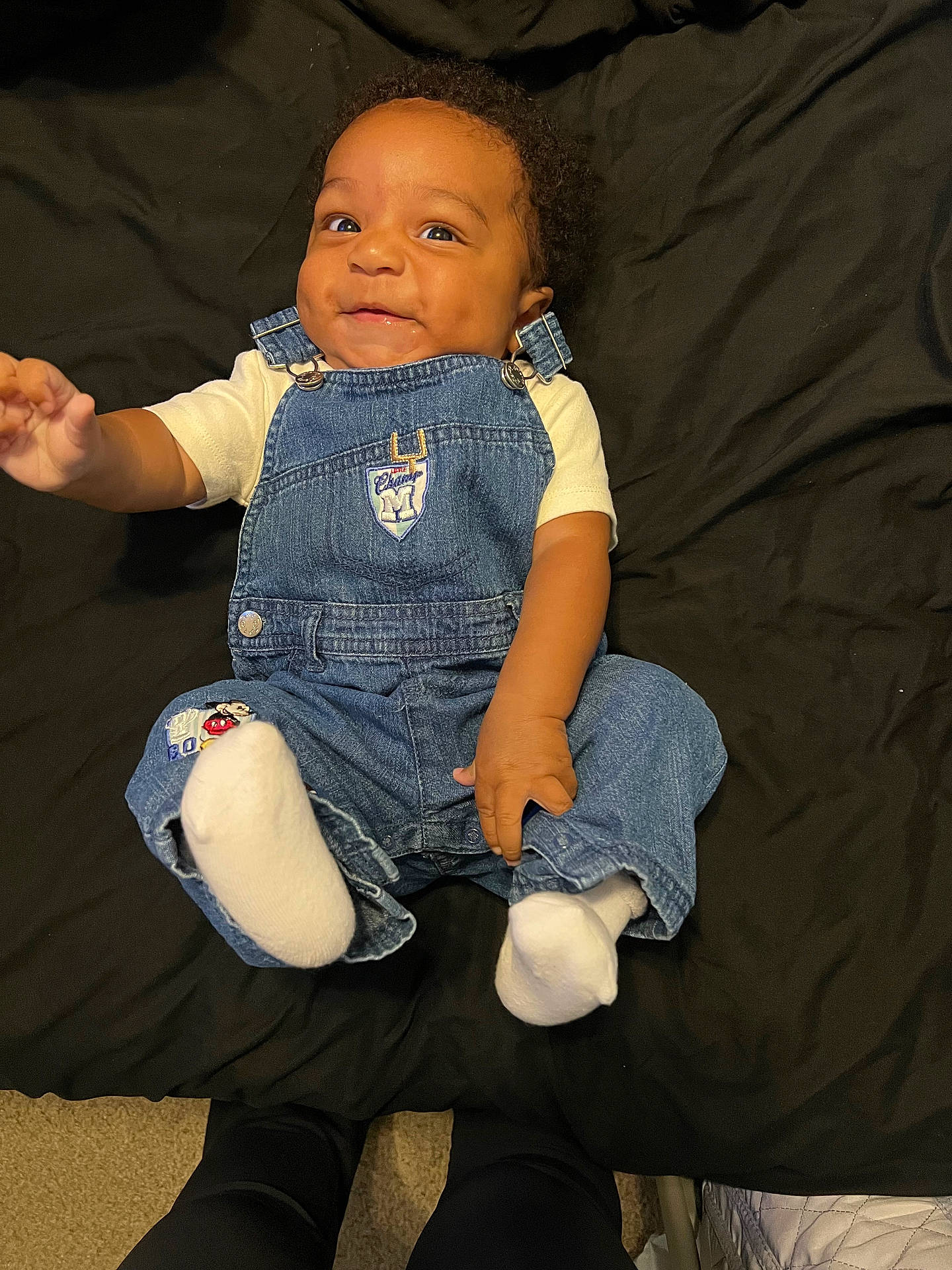 Jaxson is registered to the contest to win money with this photo: baby, black_blanket, casual, child, clothing, cute, denim_overalls, dimples, feet, hands, happy, indoors, infant, looking_up, overalls, person, portrait, sitting, smiling, white_socks