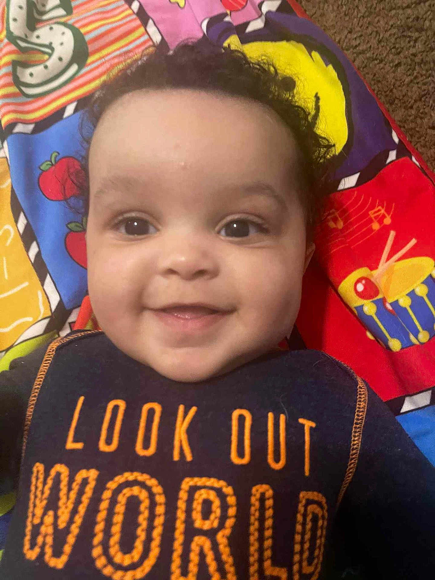 Marceau is registered to the contest to win money with this photo: baby, smile, child, curly_hair, face, clothing, text, bib, play_mat, colorful, indoor, young_child, happy, person, portrait, closeup, infant, cute, toddler, bright