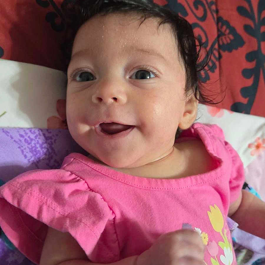 Melina joined the competition — help win amazing prizes! baby, child, smiling, pink_clothing, floral_print, lying_down, bedspread, patterned_background, happy, portrait, infant, cute, indoors, person, skin, face, head, eyes, hand, young