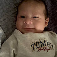 William is registered to the contest to win money with this photo: baby, infant, smile, face, clothing, sweater, text, lying_down, blanket, cute, young_child, portrait, person, apparel, indoor, soft_texture, head, eyes, fashion, comfortable