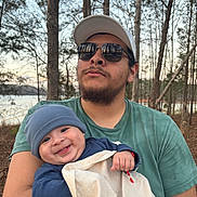 William is registered to the contest to win money with this photo: baby, adult, man, child, smile, hat, sunglasses, blanket, outdoor, forest, trees, lake, nature, person, holding, green_shirt, beard, cap, happy, daylight