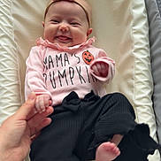 Aubrey joined the competition — help win amazing prizes! baby, infant, smiling, pink_bow, headband, pink_shirt, pumpkin_graphic, black_pants, frilled_cuffs, hand_holding, adult_hand, cushion, cream_cushion, lying_down, happy, cute, child, portrait, indoors, clothing