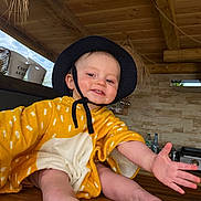 Emilien participe au concours pour gagner de l'argent avec cette photo : baby, child, smiling, hat, yellow_clothing, barefoot, wooden_table, indoor, kitchen, cupboard, wooden_ceiling, happy, person, infant, reaching_hand, face, light, casual, cute, playful