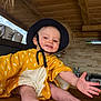 baby, child, smiling, hat, yellow_clothing, barefoot, wooden_table, indoor, kitchen, cupboard, wooden_ceiling, happy, person, infant, reaching_hand, face, light, casual, cute, playful
