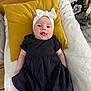 Lily a rejoint le concours — aidez-le/la à gagner de superbes lots ! baby, infant, headband, bow, black_dress, pillow, yellow_pillow, white_bedding, tongue_out, lying_down, cute, indoors, wood_floor, baby_clothing, child, smiling, face, person, home, cozy