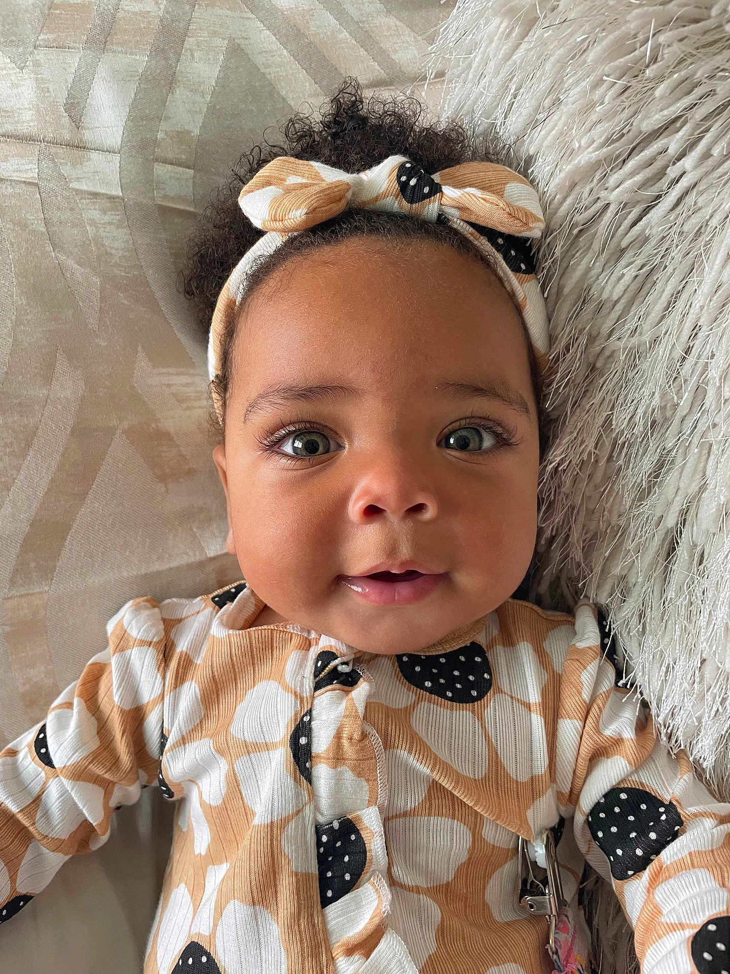Kamaya is registered to the contest to win money with this photo: baby, infant, face, headband, curly_hair, smile, eyes, clothing, pattern, cushion, blanket, portrait, closeup, cute, child, indoors, soft_texture, happy, person, lying_down
