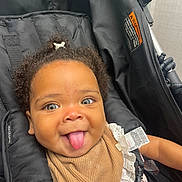 Kamaya joined the competition — help win amazing prizes! baby, child, stroller, curly_hair, tongue_out, smiling, bib, white_lace, brown_clothing, black_stroller, cute, playful, infant, seatbelt, happy, portrait, indoors, closeup, person, young_child