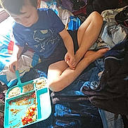 Elijah is registered to the contest to win money with this photo: child, boy, bed, blanket, stuffed_toy, teddy_bear, food, plate, fork, indoor, casual_clothing, shorts, tshirt, natural_light, window, wooden_wall, relaxed, eating, cross_legged, home