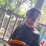 Atreyu is registered to the contest to win money with this photo: child, boy, outdoor, porch, wooden_floor, trees, playhouse, toy, camo_jacket, plaid_shorts, holding, sunlight, daytime, fence, play, smile, person, casual_clothing, nature, backyard