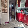 dog, small_dog, kitchen, red_cabinets, tiled_floor, doorway, dog_bowls, water_bottle, coffee_machine, paper_towel, utensils, countertop, indoor, brick_wall, chair, radiator, hallway, picture_frame, door, natural_light