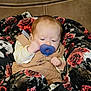 baby, pacifier, blanket, floral_pattern, couch, brown, beige_outfit, infant, child, face, hand, comfort, soft_texture, indoor, portrait, clothing, cute, resting, young_child, warm_colors
