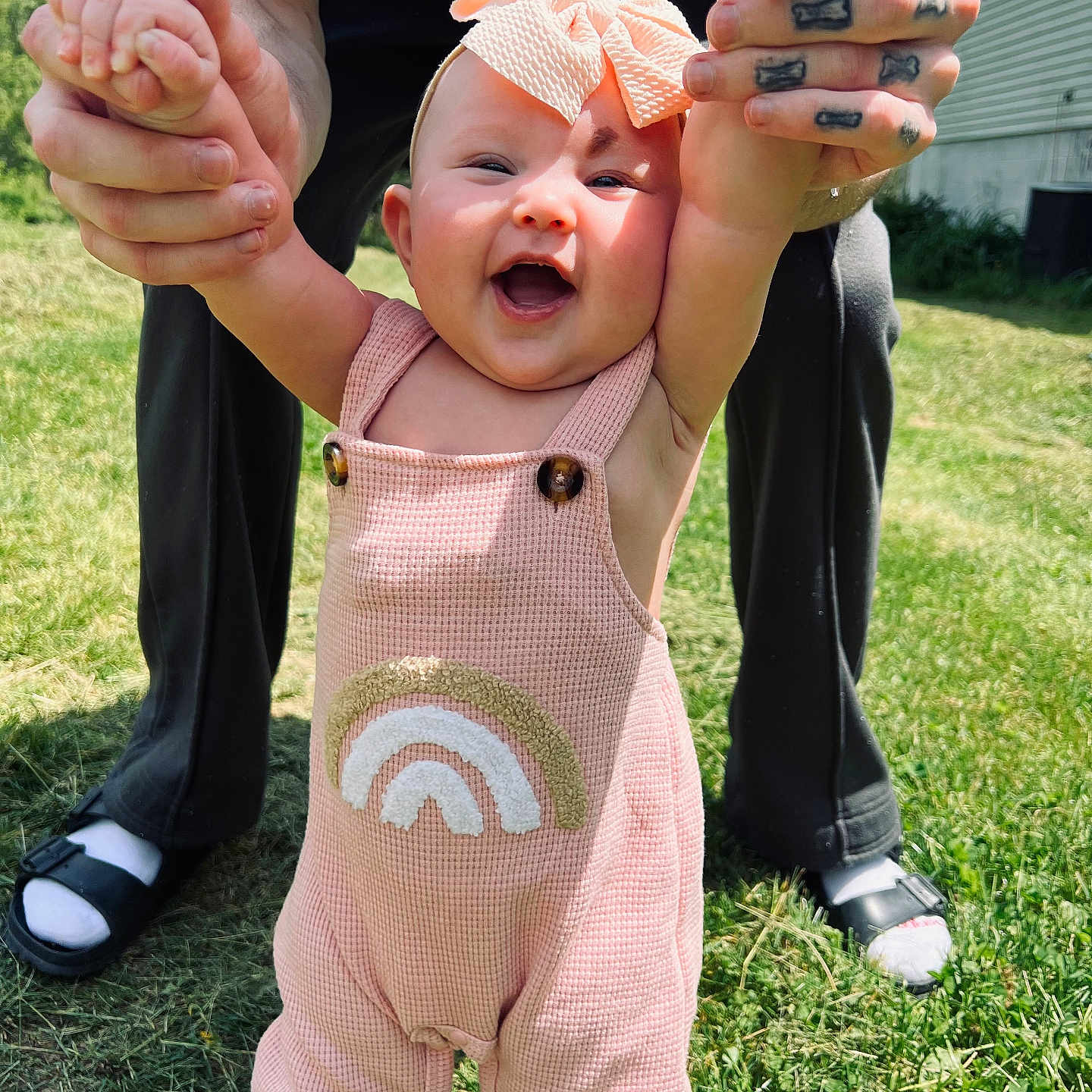 Briley is registered to the contest to win money with this photo: baby, child, smiling, happy, outdoor, grass, sunlight, headband, pink_outfit, rainbow_design, adult_hands, tattoo, holding_hands, cute, playful, person, footwear, casual_clothing, nature, daylight