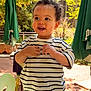 toddler, child, smiling, striped_shirt, curly_hair, outdoor, sunlight, green_umbrella, trees, fence, jeans, happy, person, daylight, playing, nature, park, young_child, casual_clothing, portrait