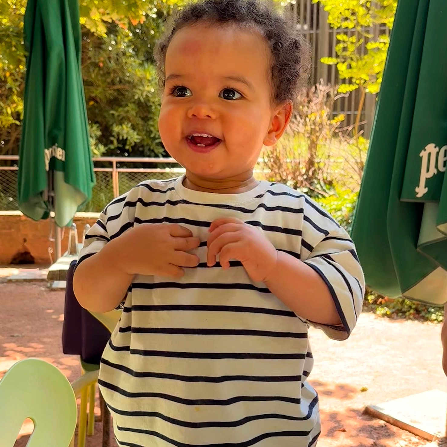 Vasco a rejoint le concours — aidez-le/la à gagner de superbes lots ! casual_clothing, child, curly_hair, daylight, fence, green_umbrella, happy, jeans, nature, outdoor, park, person, playing, portrait, smiling, striped_shirt, sunlight, toddler, trees, young_child