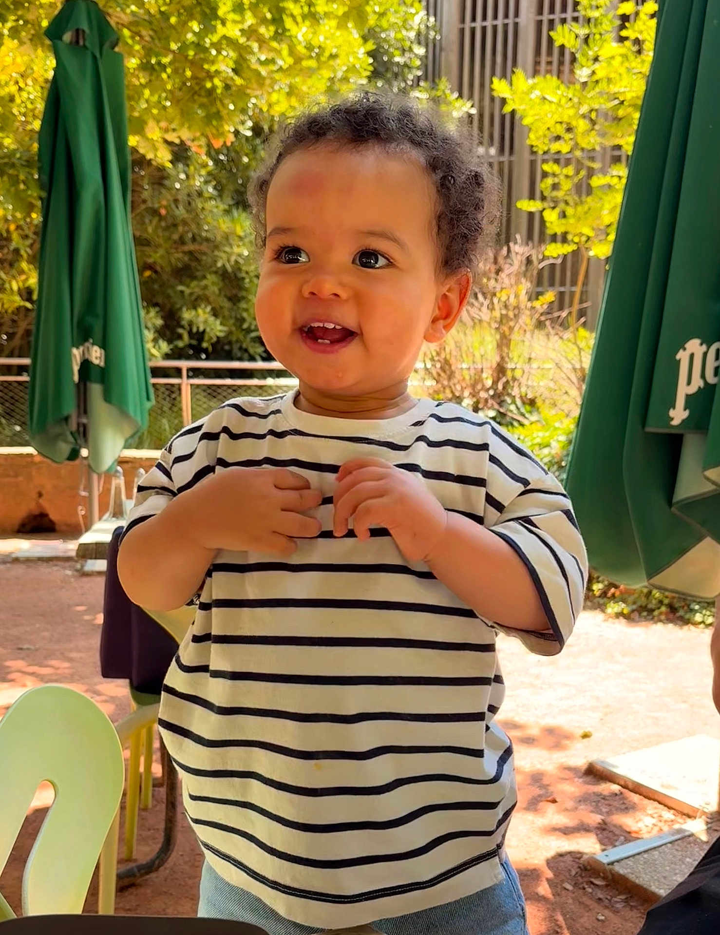 Vasco a rejoint le concours — aidez-le/la à gagner de superbes lots ! toddler, child, smiling, striped_shirt, curly_hair, outdoor, sunlight, green_umbrella, trees, fence, jeans, happy, person, daylight, playing, nature, park, young_child, casual_clothing, portrait