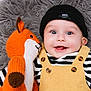 baby, child, smiling, knitted_toy, fox, beanie, yellow_overalls, striped_shirt, happy, cute, soft_blanket, portrait, indoor, close_up, playful, infant, toy, cozy, button, expression