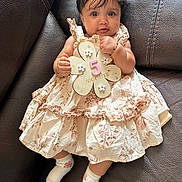 Safia is registered to the contest to win money with this photo: baby, bracelet, child, couch, cute, dress, floral_pattern, flower_shape, hair_clip, hand, holding, indoors, infant, leather, portrait, sign, sitting, smiling, socks, young_child