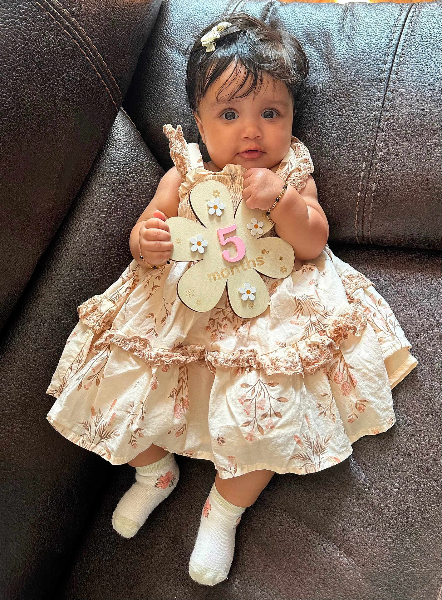 Safia is registered to the contest to win money with this photo: baby, bracelet, child, couch, cute, dress, floral_pattern, flower_shape, hair_clip, hand, holding, indoors, infant, leather, portrait, sign, sitting, smiling, socks, young_child