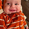 baby, infant, child, smiling, happy, portrait, close_up, face, eyes, mouth, orange_clothing, striped_clothing, hoodie, fabric, cozy, indoor, warm_lighting, lying_down, photography, adorable
