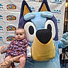 baby, child, infant, mascot, cartoon_dog, blue_dog, portrait, event, backdrop, childrens_event, hospital, purple_clothing, onesie, smiling, sitting, indoors, photography, tattooed_arm, plush_costume, colorful