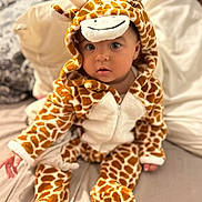Norah a rejoint le concours — aidez-le/la à gagner de superbes lots ! baby, child, giraffe_costume, onesie, bed, pillow, blanket, indoor, cute, infant, soft_toy_style, animal_print, childrens_clothing, portrait, sitting, looking_at_camera, cozy, warm, plush, adorable