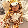 baby, child, giraffe_costume, onesie, bed, pillow, blanket, indoor, cute, infant, soft_toy_style, animal_print, childrens_clothing, portrait, sitting, looking_at_camera, cozy, warm, plush, adorable