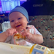 Taybian is registered to the contest to win money with this photo: baby, high_chair, bib, food, messy, smiling, hat, indoor, feeding, hands, face, child, person, jar, table, expression, cute, clothing, furniture, happy