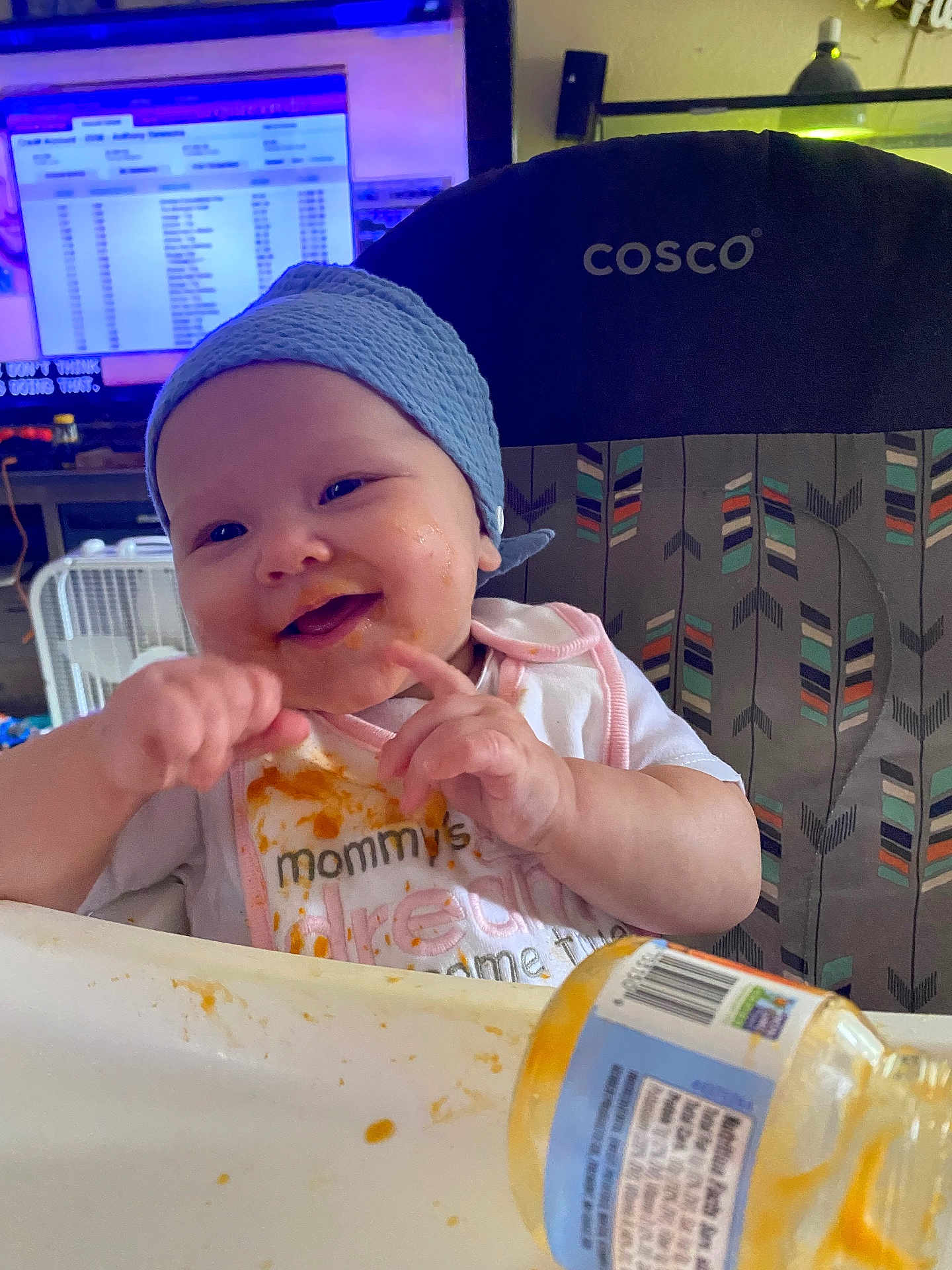 Taybian is registered to the contest to win money with this photo: baby, high_chair, bib, food, messy, smiling, hat, indoor, feeding, hands, face, child, person, jar, table, expression, cute, clothing, furniture, happy