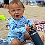 baby, child, beach, sand, water_bottle, swimsuit, shark_pattern, blanket, sitting, outdoor, sunny, smiling, happy, person, infant, summer, vacation, toy, playful, fun