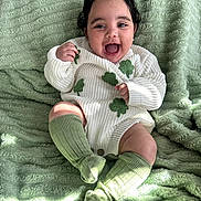 Alani is registered to the contest to win money with this photo: baby, smiling, happy, sitting, green_socks, white_sweater, clover_pattern, curly_hair, blanket, soft_texture, cozy, indoors, child, cute, portrait, infant, warm_light, expression, comfortable, playful