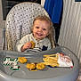 chicken_nugget, child, clothing, floor, food, french_fries, furniture, happy, high_chair, indoor, jacket, meal, person, radiator, reflection, smiling, sock, toddler, tray, window