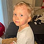 toddler, child, face, blue_eyes, blond_hair, messy_mouth, sitting, white_shirt, brown_pants, indoor, couch, curious, casual, home, window, blanket, toy, furniture, light, portrait