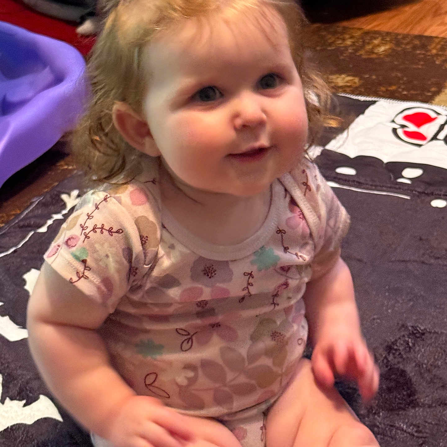 Chloe is registered to the contest to win money with this photo: baby, child, curly_hair, bow, sitting, blanket, indoor, floor, toy, patterned_clothing, cute, chubby_cheeks, infant, smile, person, head, face, legs, furniture, carpet