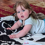 Chloe joined the competition — help win amazing prizes! baby, child, infant, curly_hair, blanket, floor, rug, remote_control, indoor, crawling, cute, person, home, wood_floor, patterned_blanket, white_clothing, blue_eyes, cheeks, curious, laying_down