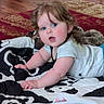 baby, child, infant, curly_hair, blanket, floor, rug, remote_control, indoor, crawling, cute, person, home, wood_floor, patterned_blanket, white_clothing, blue_eyes, cheeks, curious, laying_down