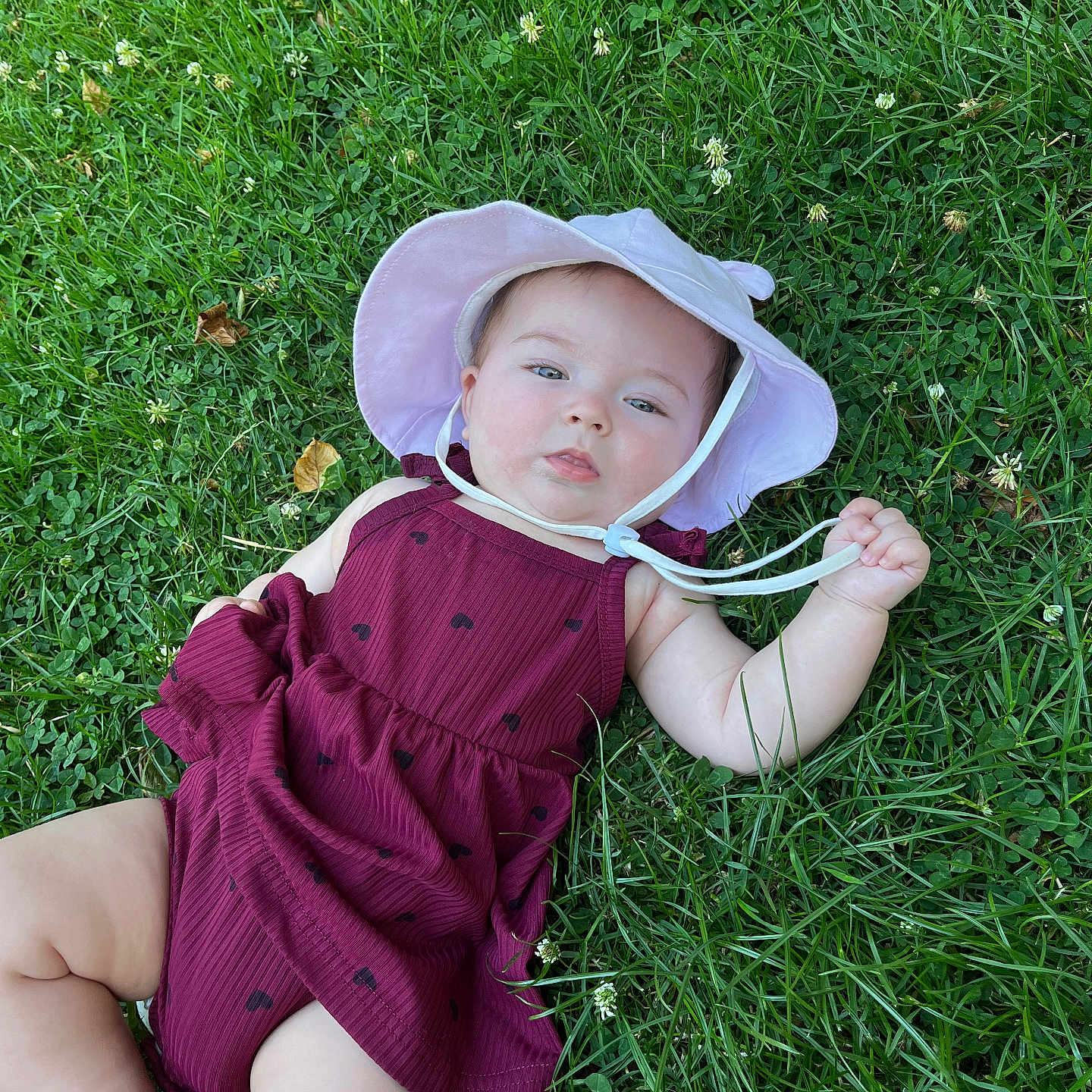 Athéna participe au concours pour gagner de l'argent avec cette photo : baby, child, cute, daylight, dress, expression, face, grass, greenery, hand, infant, legs, lying_down, maroon, nature, outdoor, playful, skin, summer, sunhat