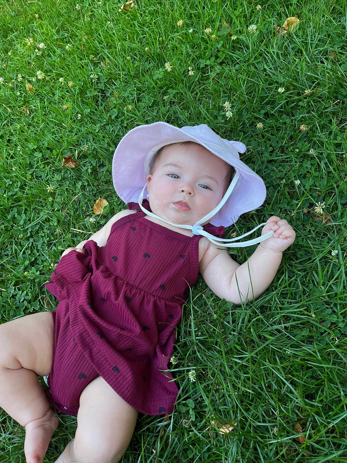 Athéna participe au concours pour gagner de l'argent avec cette photo : baby, child, grass, outdoor, sunhat, dress, maroon, greenery, infant, cute, nature, lying_down, summer, hand, legs, skin, face, expression, playful, daylight