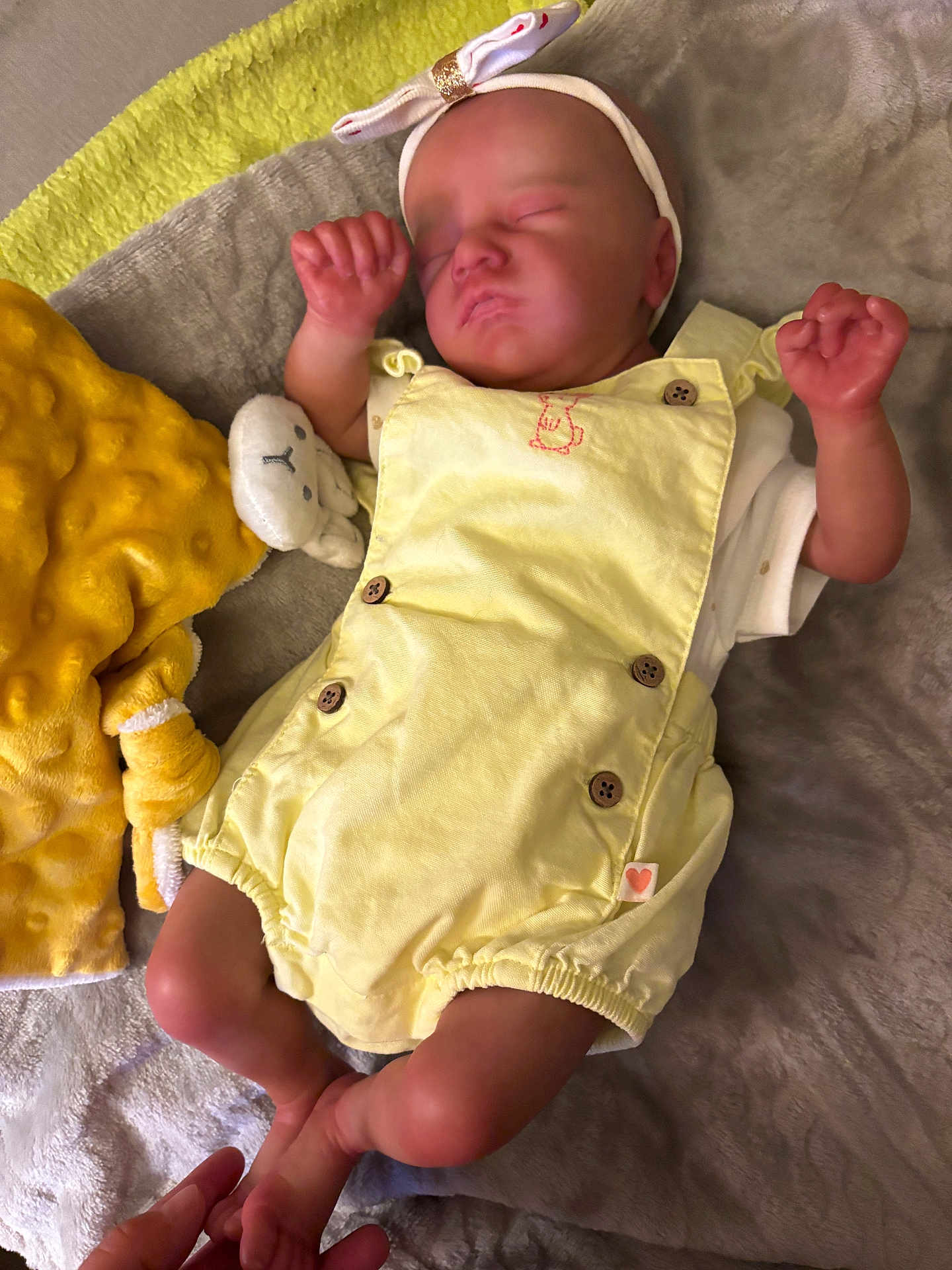 Manon a rejoint le concours — aidez-le/la à gagner de superbes lots ! baby, sleeping, yellow_romper, headband, bow, blanket, plush_toy, hand, infant, soft_texture, clothing, cute, peaceful, newborn, resting, indoors, person, skin, foot, hand_holding