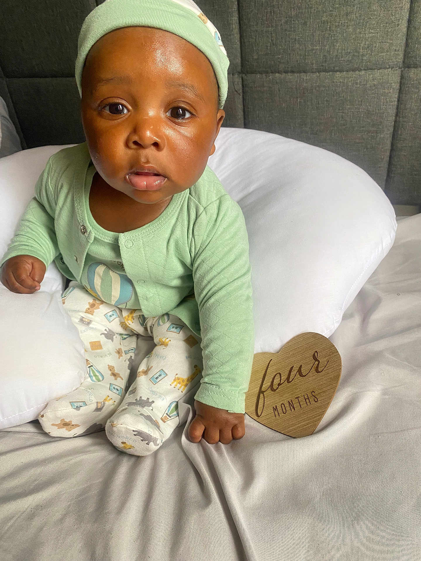 Nazir joined the competition — help win amazing prizes! baby, infant, child, pajamas, green_clothing, headwear, cushion, bed, white_bedding, wooden_sign, heart_shaped, four_months, portrait, indoor, cute, skin, face, eyes, hand, sitting