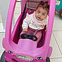 toddler, child, toy_car, pink, indoor, floor_tiles, steering_wheel, curly_hair, clothing, sitting, play, cute, face, eyes, expression, vehicle, small_wheels, plastic, buckle, shopping_cart