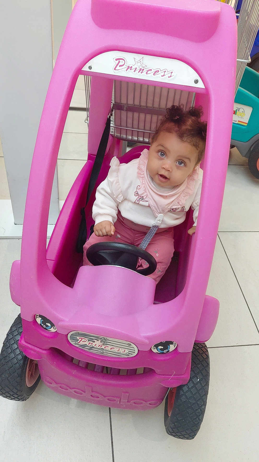 Talya a rejoint le concours — aidez-le/la à gagner de superbes lots ! toddler, child, toy_car, pink, indoor, floor_tiles, steering_wheel, curly_hair, clothing, sitting, play, cute, face, eyes, expression, vehicle, small_wheels, plastic, buckle, shopping_cart