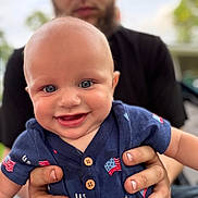 John is registered to the contest to win money with this photo: adult, baby, blue_eyes, blurred_background, buttons, casual_clothing, child, cute, face, flag_pattern, hands, happy, holding, infant, outdoor, person, portrait, shirt, skin, smiling