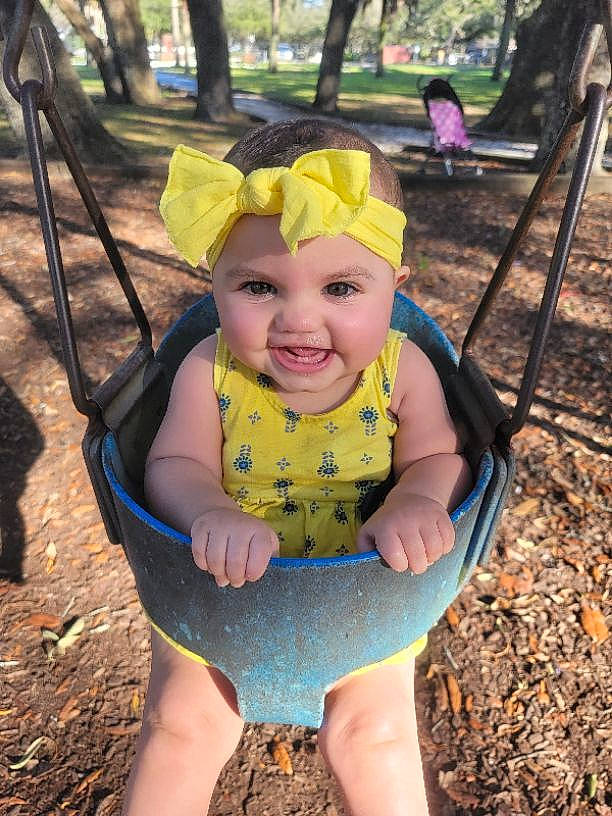 Davina is registered to the contest to win money with this photo: baby, chair, city, community, eye, fun, grass, happy, headwear, human_settlement, joy, leaf, leisure, outdoor_play_equipment, person, personal_protective_equipment, plant, recreation, smile, swing