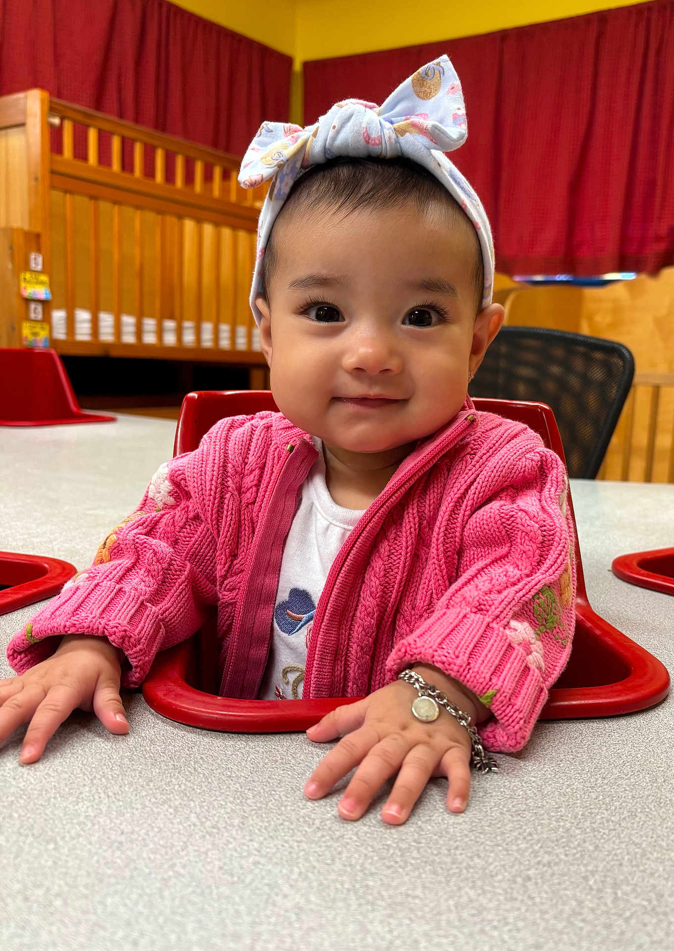 Arely joined the competition — help win amazing prizes! baby, child, smiling, headband, pink_sweater, bracelet, table, chair, indoor, crib, curtains, wooden_furniture, person, portrait, cute, happy, toddler, hands, face, cozy